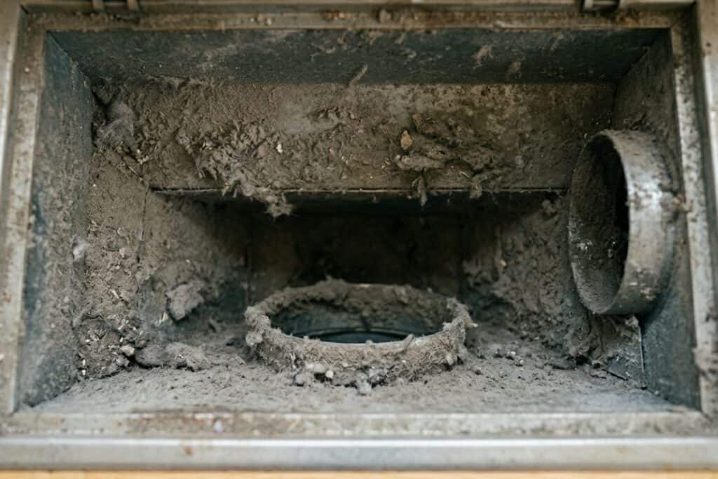 A close-up, top-down view looking into a rectangular metal floor air duct opening coated with a thick layer of grey dust, debris, and lint buildup.